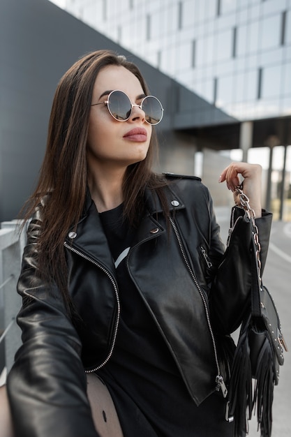 Moda stylowa piękna modelka kobieta w okrągłych okularach przeciwsłonecznych w stylu vintage z modną skórzaną kurtką i czarną sukienką ze stylową torebką spaceruje po mieście
