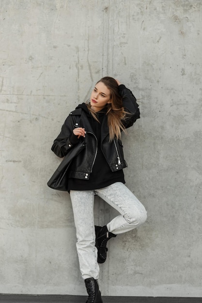 Moda urocza nastolatka kobieta model hipster w modnych ubraniach ze skórzaną kurtką czarna bluza z kapturem dżinsy i buty ze skórzaną modną torbą i pozami w pobliżu betonowej ściany na ulicy