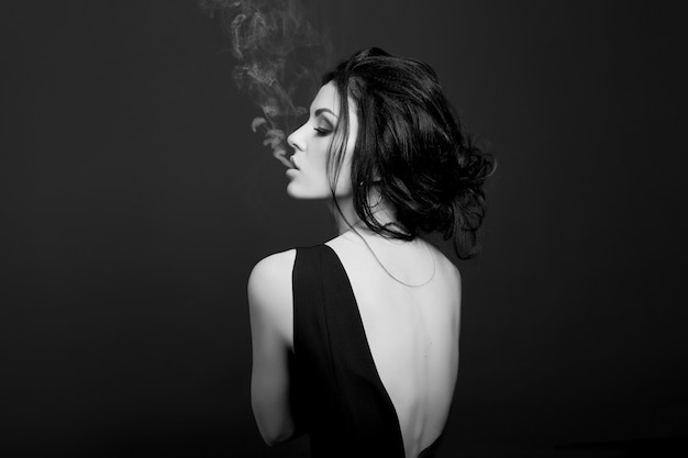 Moda uroda makijaż kosmetyki brunetka dama pali na ciemnym tle w czarnej sukience, biały dym z ust. Erotyczne dziewczyny, czarno-białe