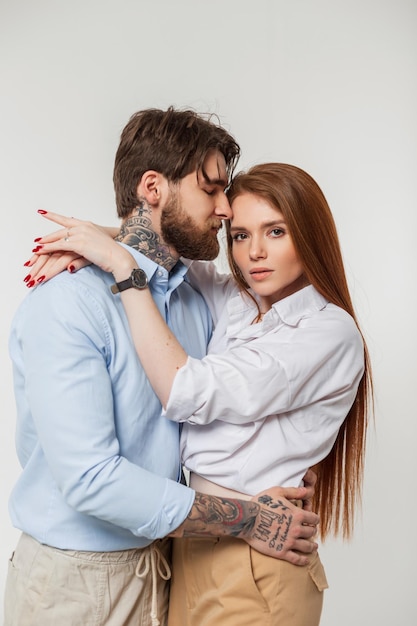 Moda uroda para piękna ruda kobieta i przystojny hipster mężczyzna z brodą i tatuażami stoi i przytula się na białym tle w studio