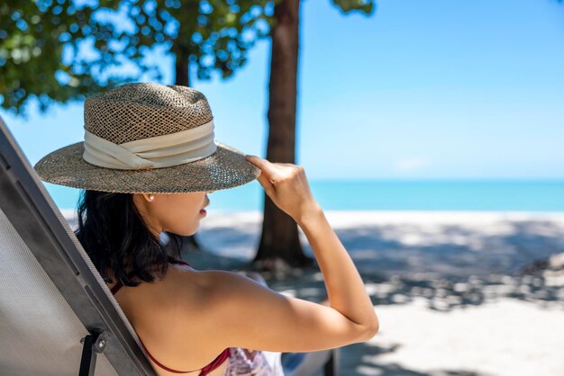 Model Azjatycka kobieta ubrana w czerwony strój kąpielowy i słomkowy kapelusz siedzi w słońcu nad morzem Piękna dziewczyna relaksuje