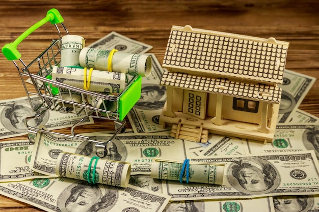Model domu i mały wózek na zakupy z rolkami dolarów na stosie banknotów stodolarowych na drewnianym tle Inwestycja w nieruchomość Kredyt hipoteczny dom hipoteczny koncepcja nieruchomości