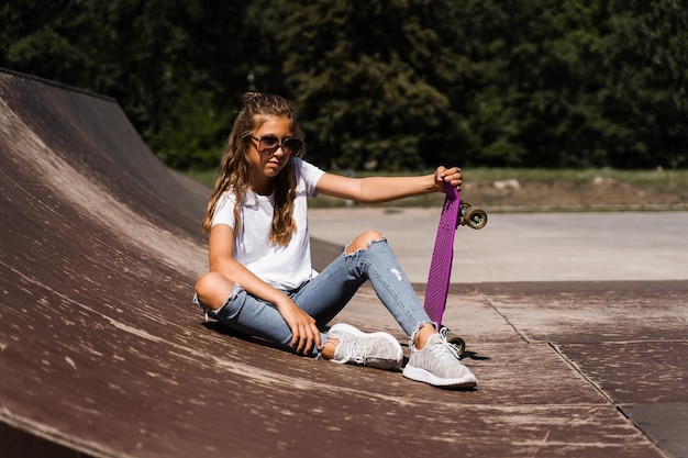 Model dziecka w okularach siedzi i pozuje z tablicą grosza na skate placu zabaw Aktywna dziewczynka z ekstremalnym stylem życia na rampie sportowej