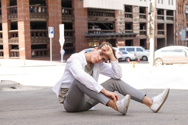 Model Facet Ze Stylową Fryzurą Pozuje Na Zewnątrz W Białej Koszuli I Szarych Spodniach. Modna Fryzura Spoczywa W Pobliżu Nowoczesnego Centrum Biznesowego. Atrakcyjny Facet Na Ulicy W Letni Dzień.