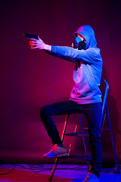 Model młody mężczyzna pozuje w studio w modnym świetle neonowym w sportowym garniturze, kapturze i masce, trzymając pistolet.