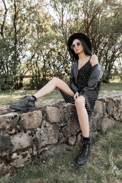 Model nowoczesny fajny kobieta z seksownymi nogami w modnych pięknych czarnych ubraniach w stylowym kapeluszu vintage w butach w okularach przeciwsłonecznych opiera się na kamieniach w parku w słoneczny wiosenny dzień. Moda dziewczyna cieszy się na zewnątrz.