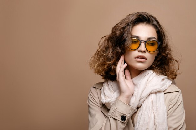 Modelka piękna z czystą skórą i kręconymi włosami w beżowym płaszczu z szalikiem na beżowej ścianie, modelka w modnych okularach