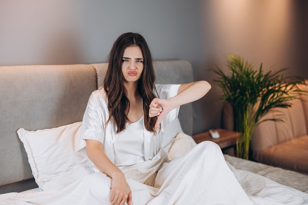 Modelka w białej jedwabnej piżamie pokazujący niechęć. piękna kobieta ze smutną twarzą siedzącą na łóżku pokazuje niechęć.