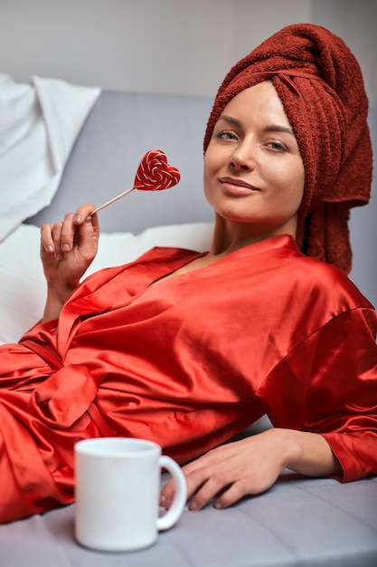 Modeluj w czerwonym szlafroku i czerwonym ręczniku na kanapie z kubkiem kawy i lizakiem. Pojęcie mody, gra kolorów, minimalizm. Odpocznij od pracy, odpocznij.