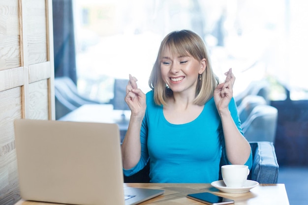 Modląc Się! Widok Z Boku Portret Freelancera życzeniowego Atrakcyjnej Młodej Dziewczyny W Niebieskiej Koszulce Siedzi W Kawiarni I Pracuje Na Laptopie, Zamknięte Oczy I Prosząc O Spełnienie Marzeń Z Uśmiechem Zębów. Wnętrz