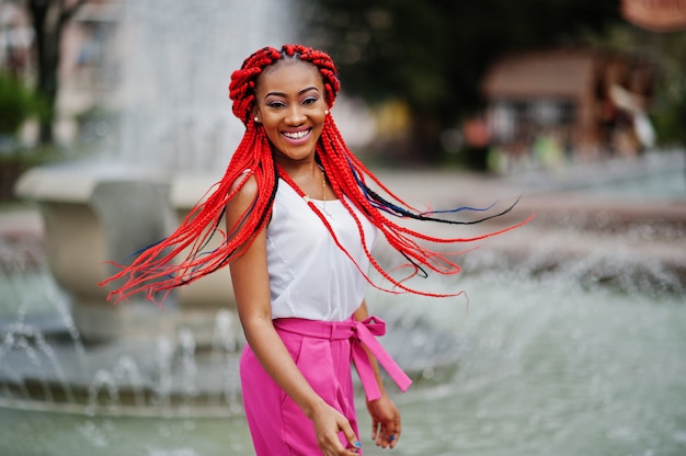 Modna amerykanin afrykańskiego pochodzenia dziewczyna przy różowymi spodniami i czerwonymi strachami pozuje na zewnątrz przeciw fontannom.