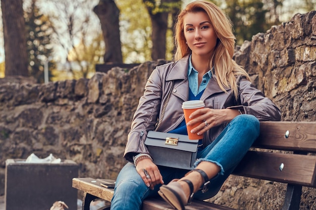 Modna Blond Kobieta Relaksuje Się Na świeżym Powietrzu, Pijąc Kawę Na Wynos, Siedząc Na ławce W Parku Miejskim.