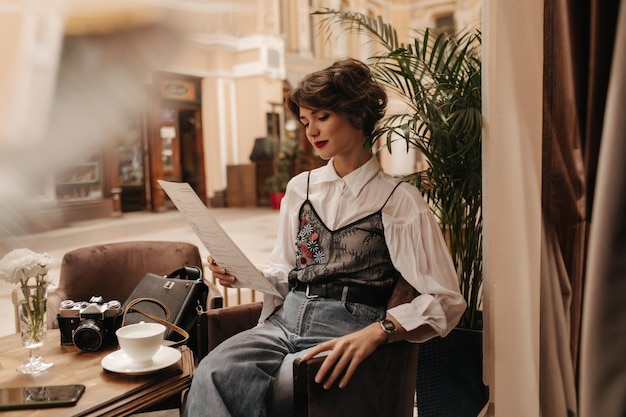 Modna dama w białej bluzce z kręconymi fryzurami menu do czytania w kawiarni Urocza dziewczyna z jasnymi ustami w dżinsach siedząca w restauracji