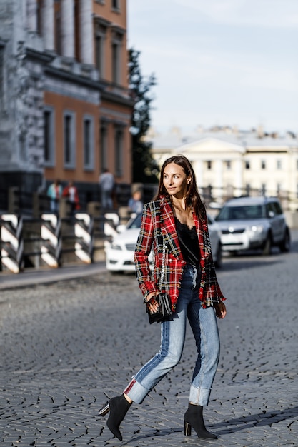 Modna dziewczyna w kraciastej kurtce i dżinsach spacerująca po mieście z czarną torebką.