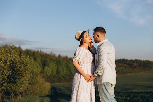 Modna i stylowa szczęśliwa kobieta w ciąży i jej mąż ubierali pastelowy biało-niebieski odcień na polu o zachodzie słońca