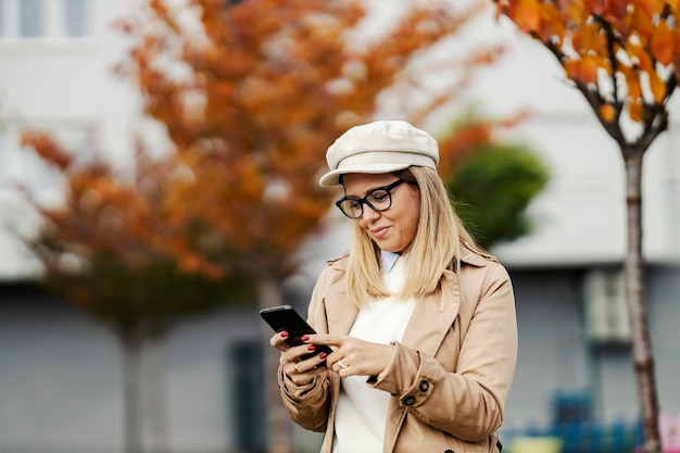 Modna kobieta stoi na ulicy miasta i uśmiecha się do telefonu