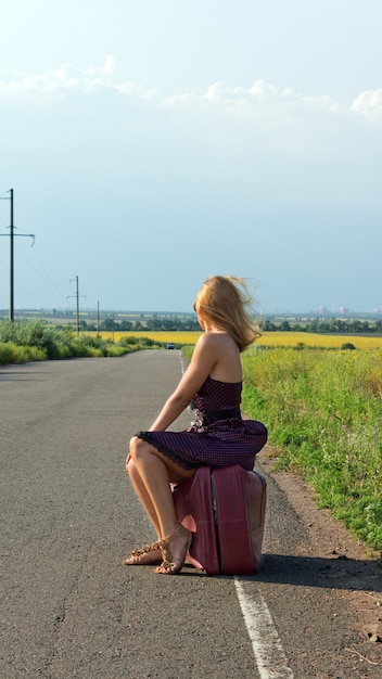 Modna kobieta w sukience i szpilkach odwrócona od kamery autostopem na poboczu drogi na wsi
