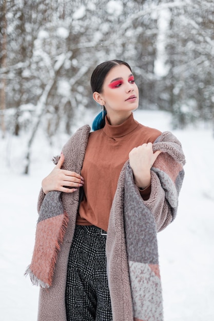 Modna młoda nastolatka z jasnym makijażem w stylowych zimowych ubraniach vintage z płaszczem, swetrem i szalikiem na śniegu w lesie