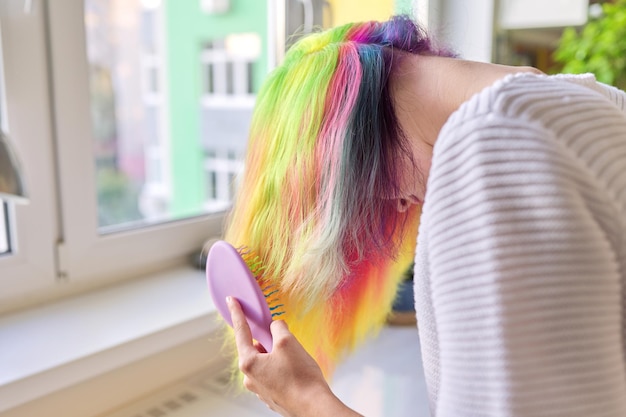Modna nastoletnia dziewczyna z modnymi tęczowymi długimi włosami czesze włosy w domu. Włosy, fryzury, kolorowanie mody, młodość i uroda