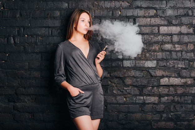 Modna piękna dziewczyna pali papierosa bez tytoniu, wydychając duże chmury dymu. Ubrany w modne ubrania.