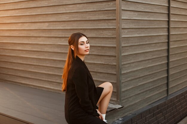 Modna, piękna, młoda modelka w czarnym stylowym płaszczu siedzi w pobliżu drewnianego domu o zachodzie słońca