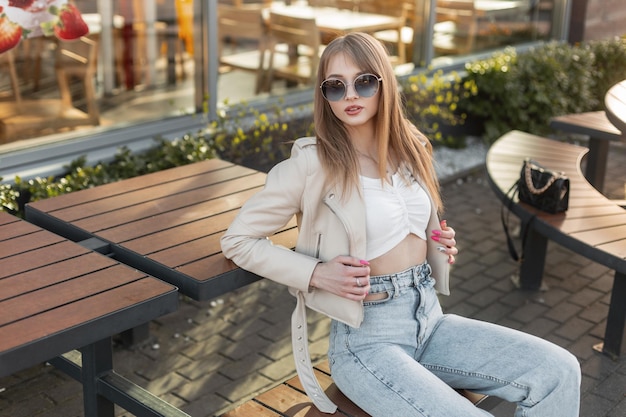 Modna piękna stylowa modelka hipster z okrągłymi okularami przeciwsłonecznymi w modnej skórzanej rockowej kurtce z topem i vintage niebieskimi dżinsami i torbą siedzącą na drewnianej ławce w pobliżu kawiarni o zachodzie słońca