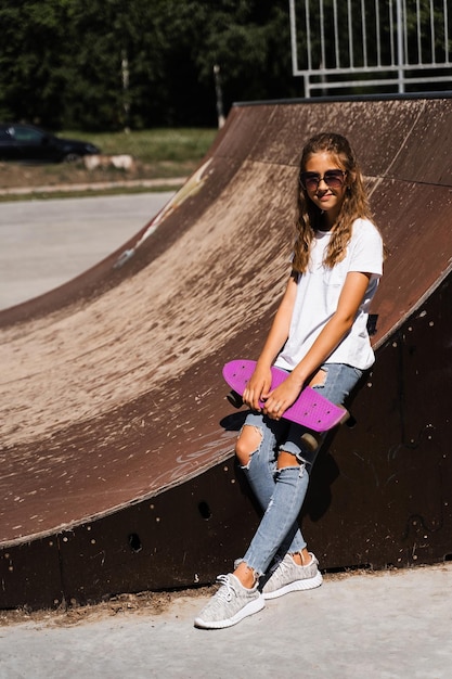 Modne dziecko dziewczynka w okularach pozuje z tablicą grosza na skate placu zabaw Aktywna nastolatka ze sprzętem sportowym na rampie sportowej Ekstremalny styl życia