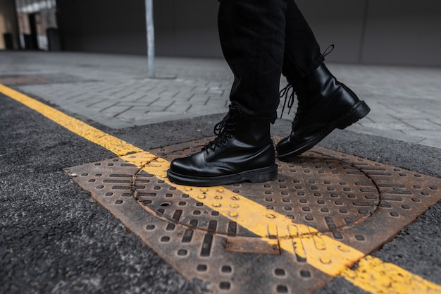 Modny młody człowiek w vintage skórzanych czarnych butach w modnych dżinsach stoi na żelaznym włazie w mieście. Zbliżenie męskich nóg w stylowych sezonowych butach. Styl uliczny. Moda młodzieżowa. Obuwie codzienne.