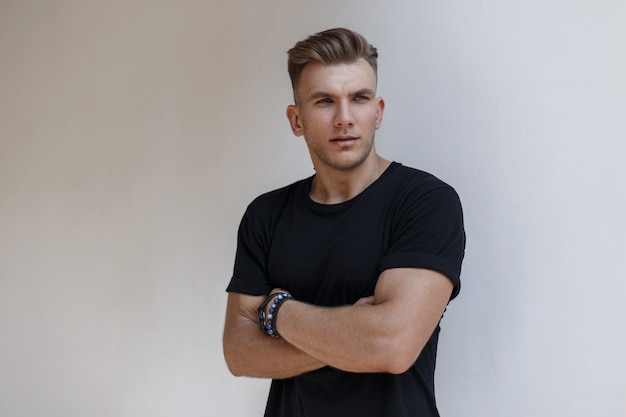 Modny przystojny amerykański model mężczyzna z fryzurą w czarnej modnej koszulce w pobliżu białej ściany