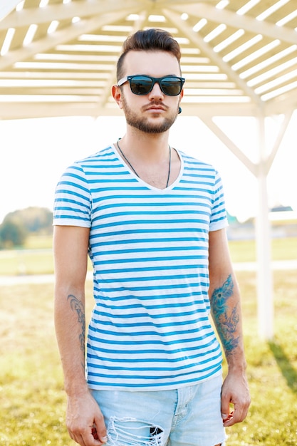 Modny przystojny mężczyzna hipster z brodą w okularach przeciwsłonecznych w modnej koszuli stoi na plaży.