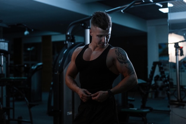 Modny przystojny młody fizycznie silny facet o muskularnym ciele w stylowej koszulce z przędzy z szortami trenuje na siłowni