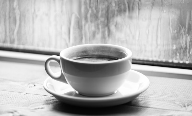 Mokre szklane okno i filiżanka gorącej kawy Jesienna pochmurna pogoda lepiej z napojem kofeinowym Ciesz się kawą w deszczowy dzień Świeżo parzona kawa w białej filiżance lub kubku na parapecie Czas kawy w deszczowy dzień
