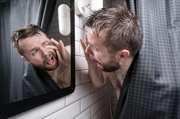 Mokry mężczyzna odczuwa dyskomfort, patrzy w lustro i próbuje wydobyć rzęsę, która dostała się do oka