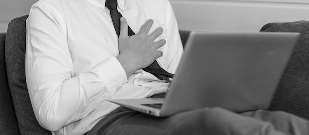 Monochromatyczne czarno-białe ujęcie nierozpoznawalnego nieznanego męskiego biznesmena pracownika w formalnym stroju biznesowym, siedzącego na wygodnej kanapie kanapie z laptopem, trzymając się za ręce na lewej piersi po zawale serca.