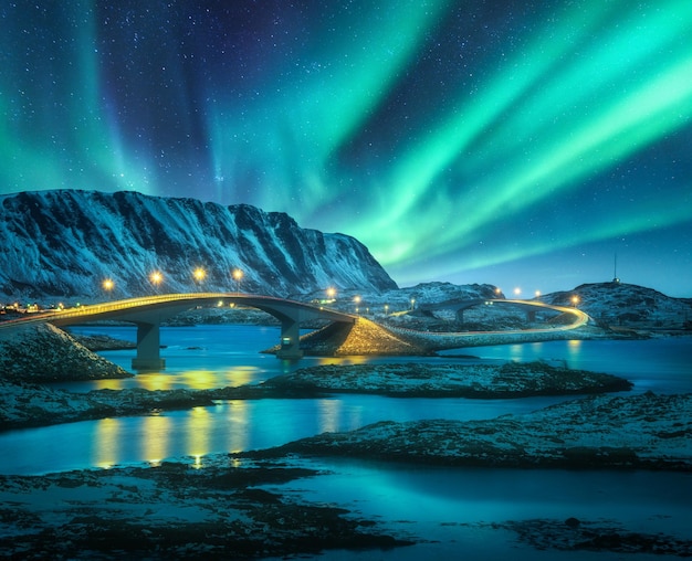 Most i zorza polarna nad ośnieżonymi górami Lofoty Norwegia Zorza polarna i odbicie w wodzie Zimowy krajobraz z rozgwieżdżonym niebem zorze polarne skały droga morze oświetlenie miasta