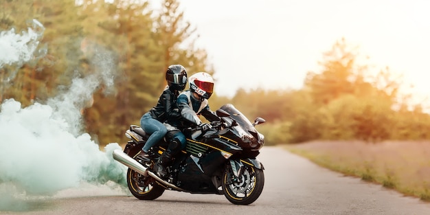 Motocyklista w ochronnym sprzęcie i kasku siedzący z dziewczyną na sportowym motocyklu na rozmytym tle z dymem, kopia przestrzeń