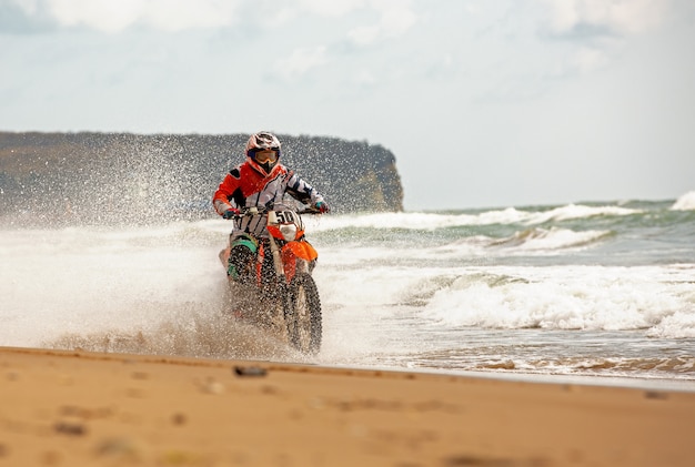 Motocyklista w stroju ochronnym jeździ motocyklem po morzu, rozpryski latają spod kół.