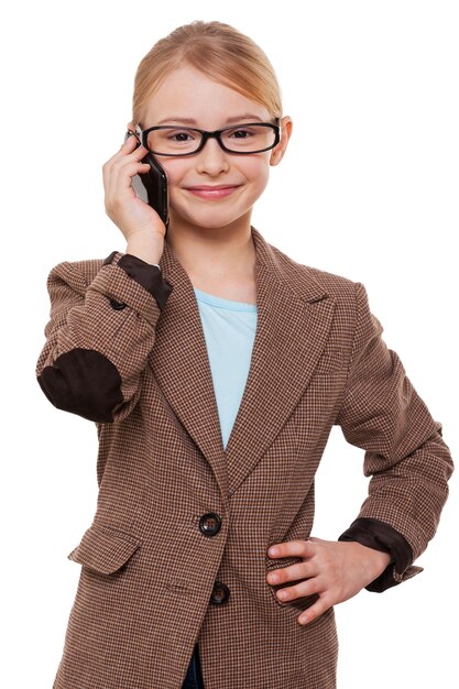 Mówiąc o biznesie. Wesoła dziewczynka w stroju formalnym rozmawia przez telefon komórkowy i trzyma rękę na biodrze, stojąc na białym tle