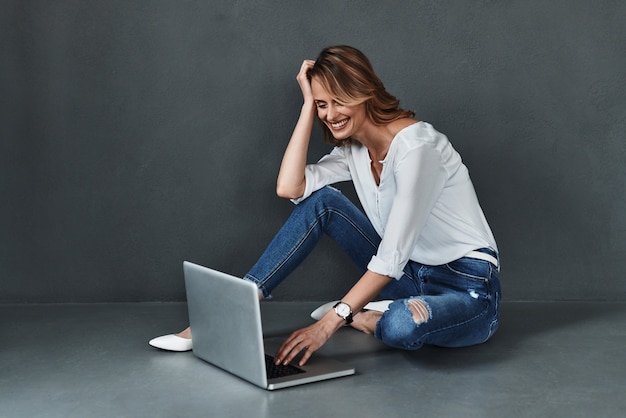 Możesz znaleźć wszystko w Internecie. Atrakcyjna młoda kobieta w stroju casual, korzystająca z komputera i uśmiechająca się siedząc na podłodze na szarym tle