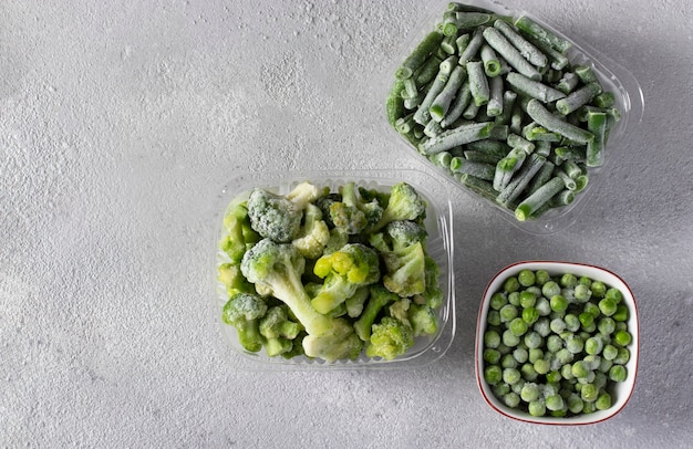 Mrożone warzywa takie jak zielony groszek, fasolka szparagowa i brokuły w pudełkach do przechowywania na jasnoszarym tle. Widok z góry, skopiuj przestrzeń