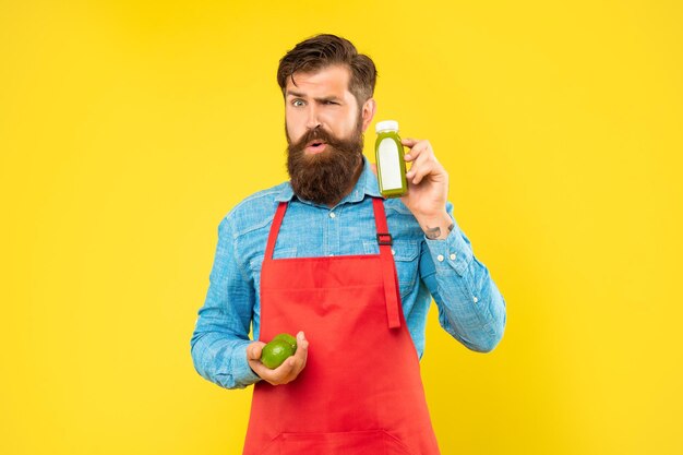 Mrugający mężczyzna w fartuchu trzymający limonki i butelkę soku na żółtym tle sklepikarz z sokiem