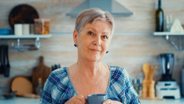 Mrużąca Starsza Kobieta Uśmiecha Się Do Kamery Trzymając Filiżankę Offee W Kuchni Podczas śniadania. Autentyczny Portret Zrelaksowanej Starszej Osoby Starszej Rano, Cieszącej Się świeżym Ciepłym Napojem. Zdrowy Uśmiech