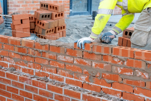 Murarz w kamizelce odblaskowej i kasku układający mur z cegły za pomocą kielni