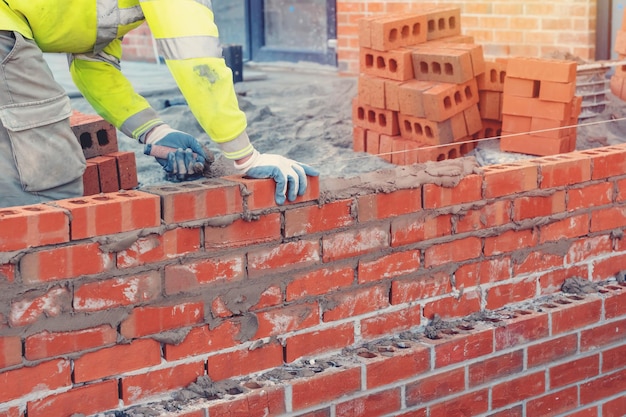 Murarz w kamizelce odblaskowej i kasku układający mur z cegły za pomocą kielni