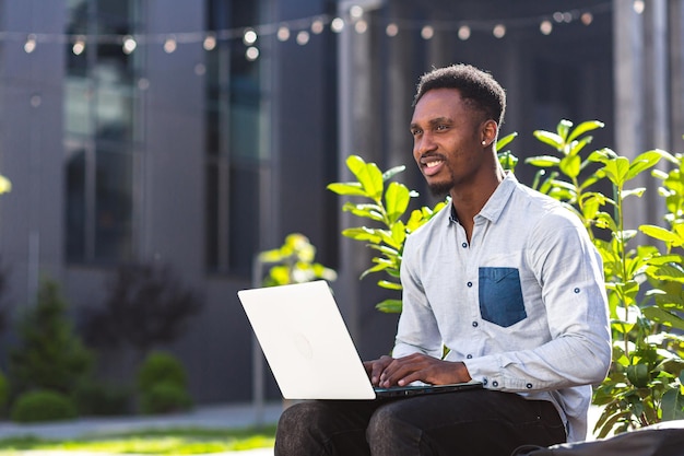 Murzyn freelancer pracujący online za pomocą laptopa siedzącego na ławce na zewnątrz nowoczesnego budynku biurowego w miejskim parku na ulicy. Szczęśliwy afroamerykanin uczeń facet w swobodnym plenerze, piszący na klawiaturze