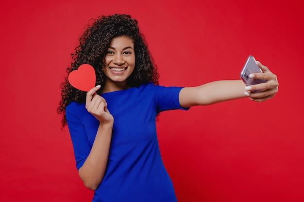 Murzynka Uśmiecha Się Selfie I Robi Selfie Z Valentine Kartą W Kształcie Serca Na Czerwonej ścianie