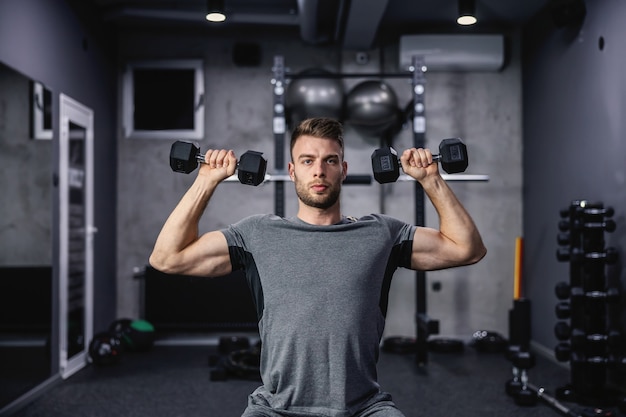 Muskularny facet podnoszenia hantle siedząc na ławce na siłowni. Młody sportowiec z hantlami podczas treningu. Silny mężczyzna podczas wysiłku fizycznego pompujący mięsień bicepsa z ciężarem