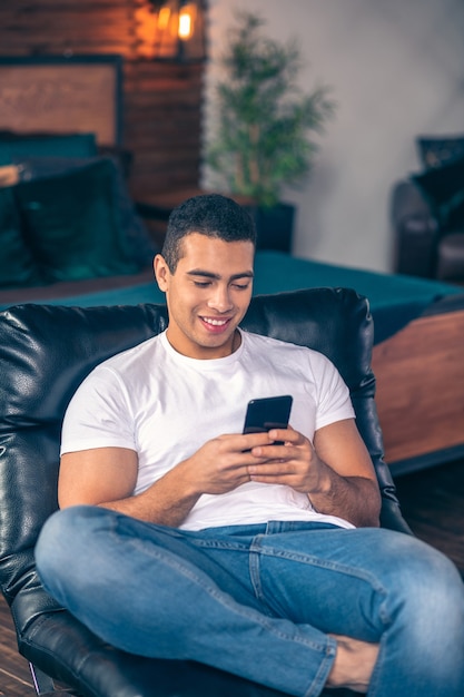Muskularny facet w białej koszulce siedzi na fotelu obok łóżka w domu, patrząc na smartfona i śmiejąc się.
