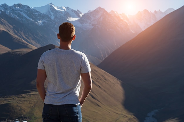 Muskularny facet w koszulce stoi i patrzy na brązowe góry skaliste z białymi ośnieżonymi szczytami z tyłu jasne jesienne światło słoneczne widok z tyłu