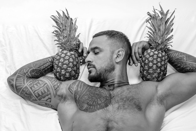 Muskularny mężczyzna trzyma owoc ananasa na bicepsie brutalny sportowiec relaks w łóżku sterydy sexy ab tatuażu
